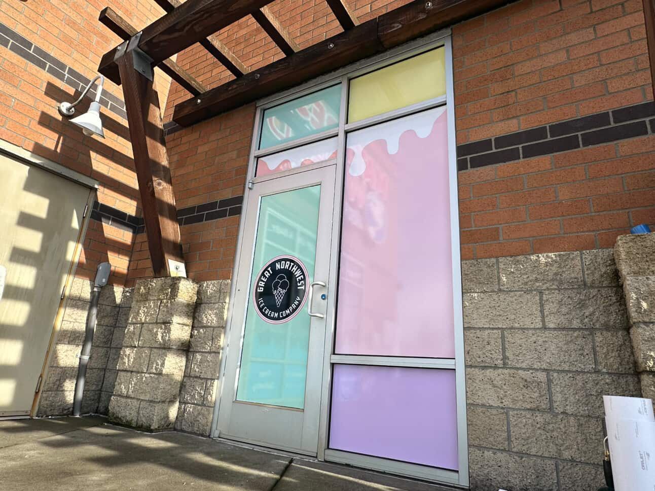 Door with colorful pastel window panels. A circular sign is on the door. Brown pergola above. Brick and stone walls.