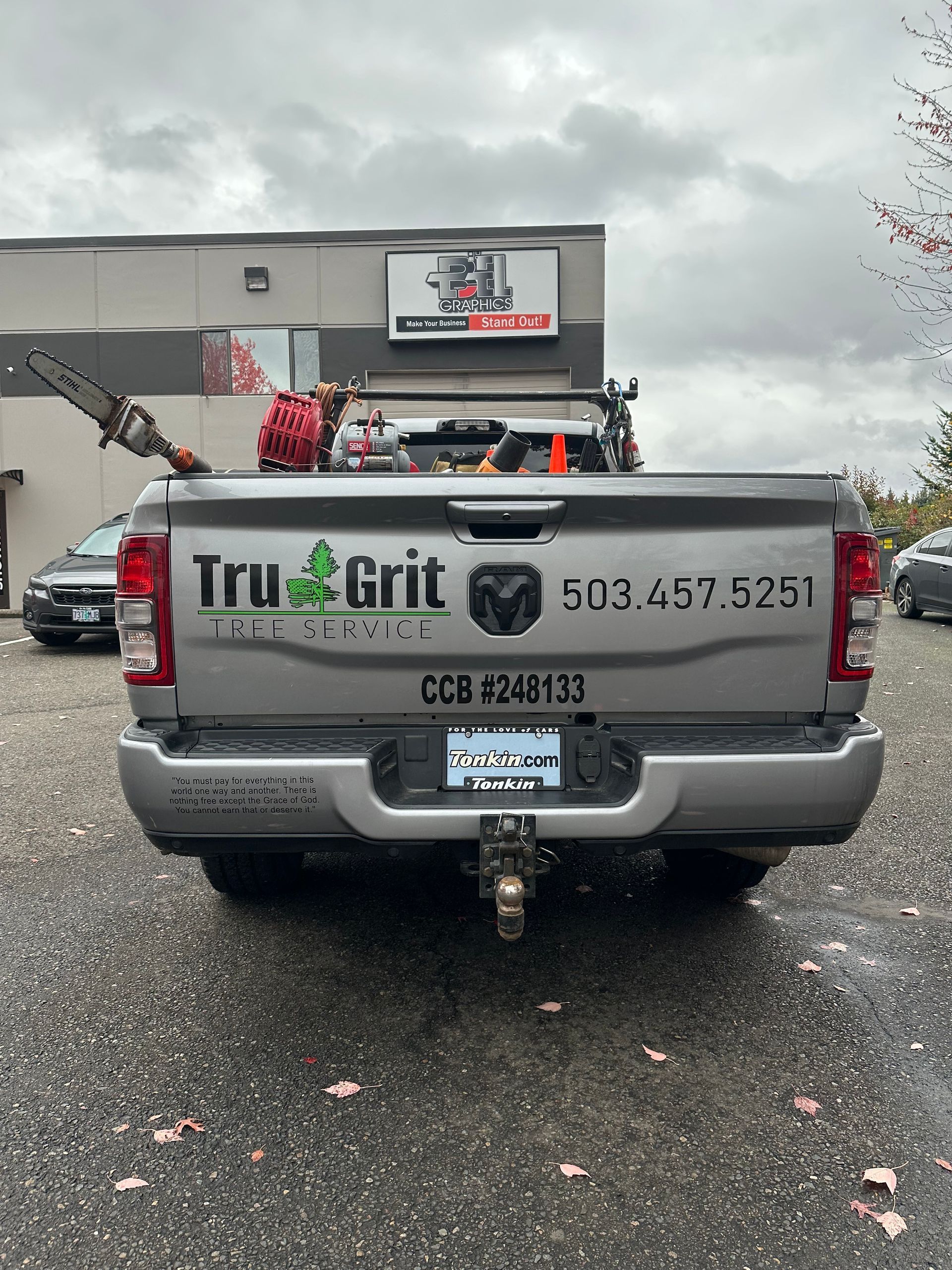 Silver Tru Grit Tree Service truck with tools, license plate, and phone number on the back, parked outdoors.