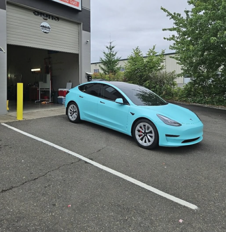 A light blue Tesla parked outside a garage, with white wheels and a black roof.