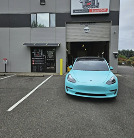 Light blue Tesla in front of a shop, possibly for car graphics. Grey building, overcast day.