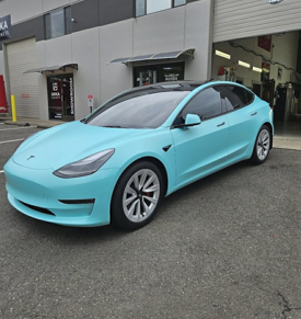 A light blue Tesla Model 3 parked in front of a building. It has a black roof and silver wheels.