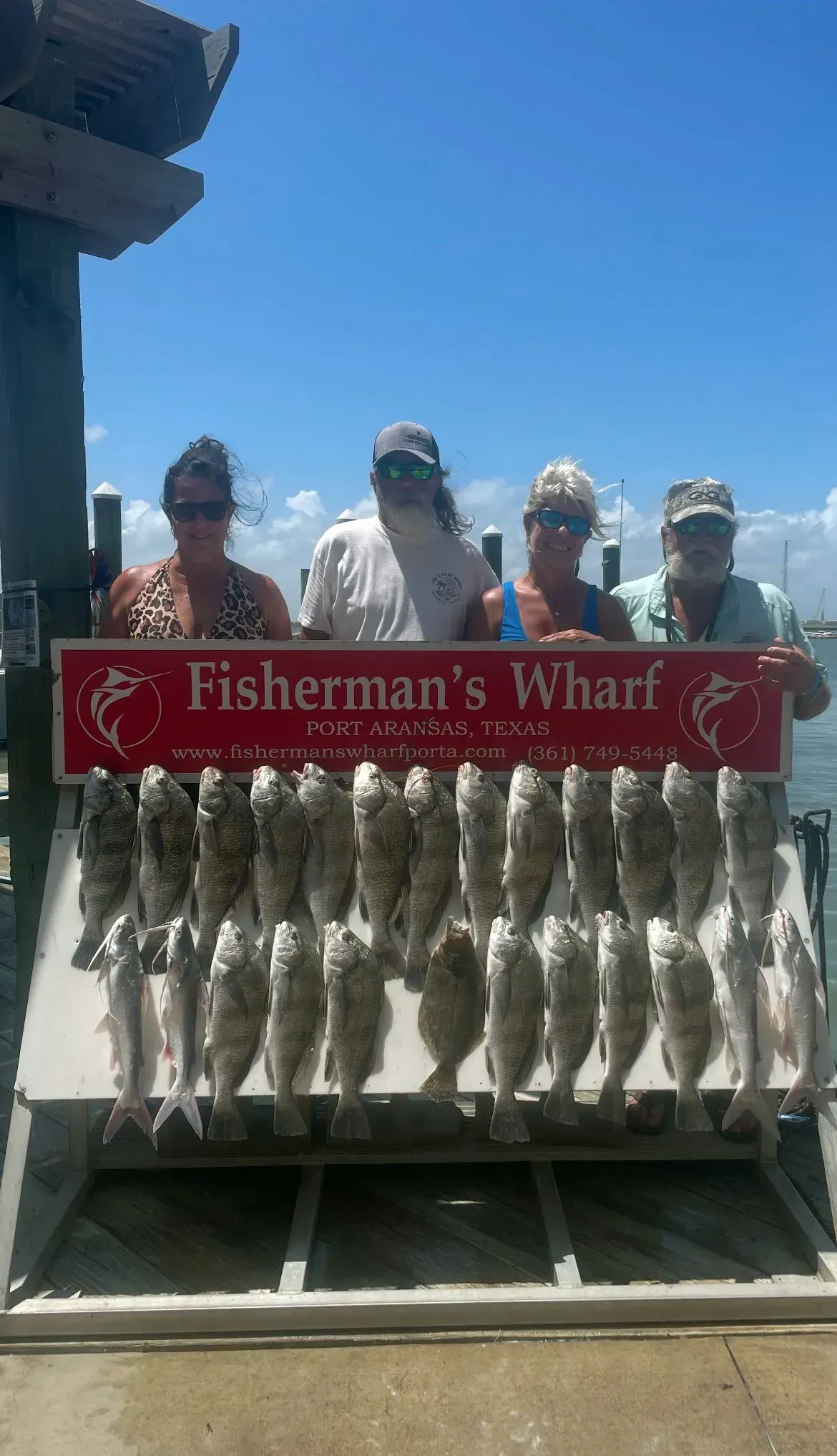 Four people pose with a large catch of fish at Fisherman's Wharf under a blue sky.