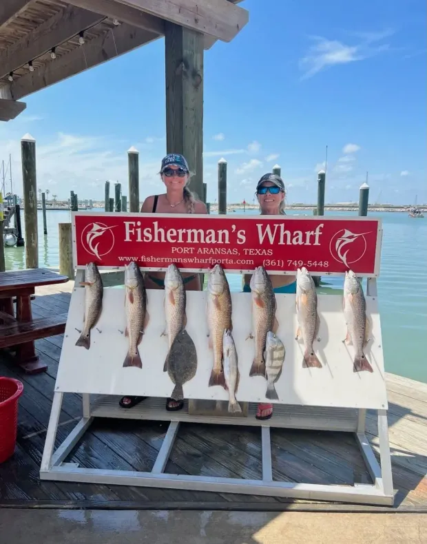 Two people posing with a fishing catch at Fisherman's Wharf, Rockport, Texas. Fish are displayed on a rack, with blue sky.