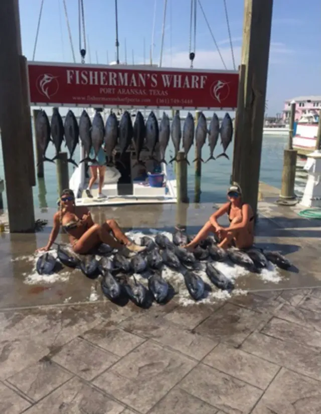Two women posing with a pile of fish on a dock, Fisherman's Wharf sign in the background, fish hanging.