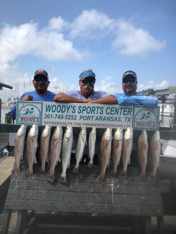 Three men display a catch of fish at Woody's Sports Center in Port Aransas, TX. Blue sky, water, and pier visible.