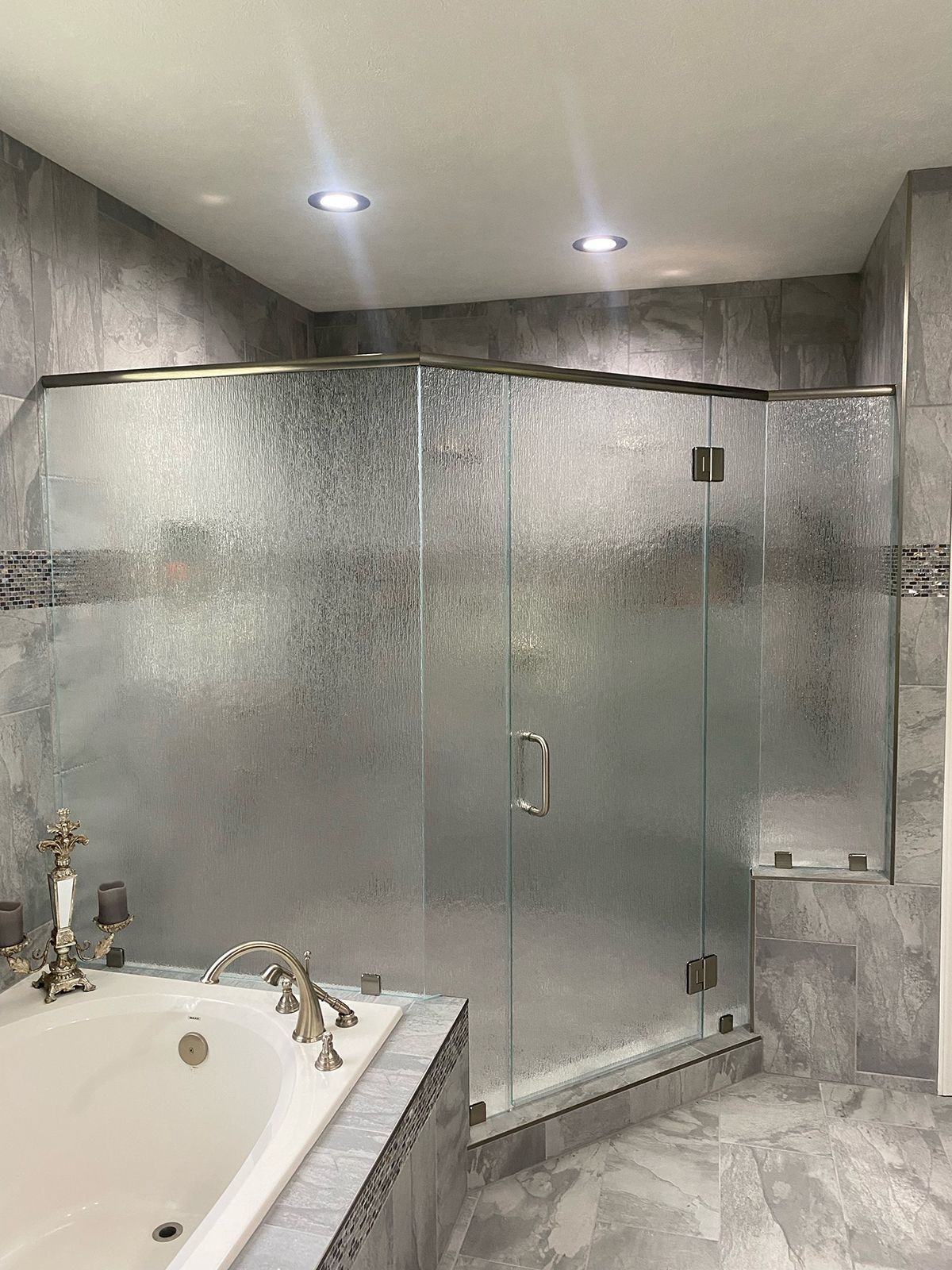 A modern glass shower enclosure with textured panels and silver hardware, situated next to a bathtub in a tiled bathroom.