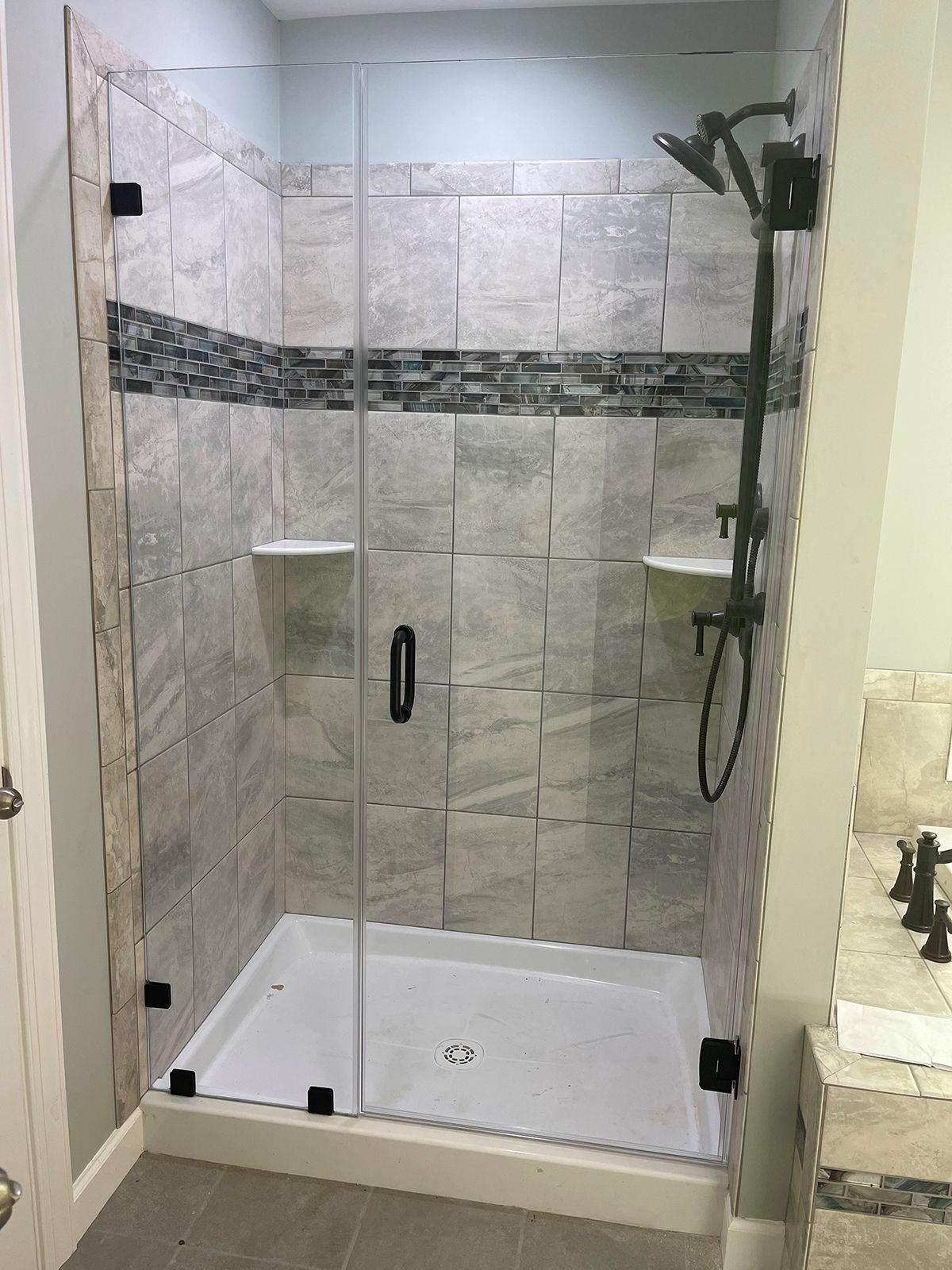 A modern walk-in shower with light grey tile walls, a dark stone accent strip, glass door, and matte black hardware.