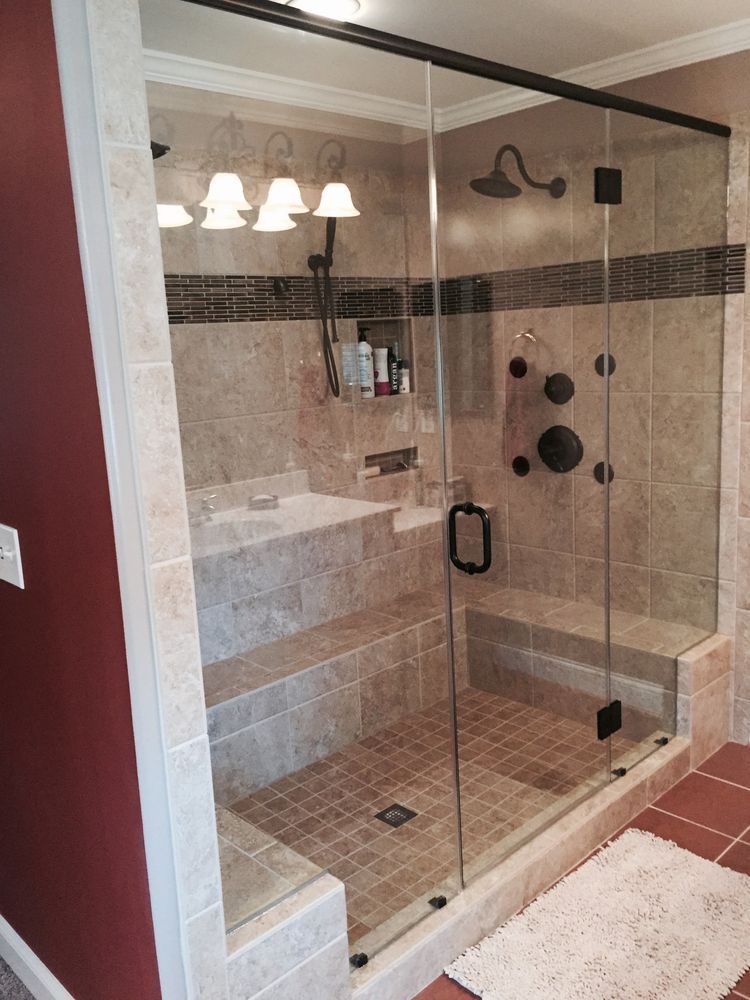 Walk-in shower with glass doors, stone tile, built-in bench, dark fixtures, and white rug.