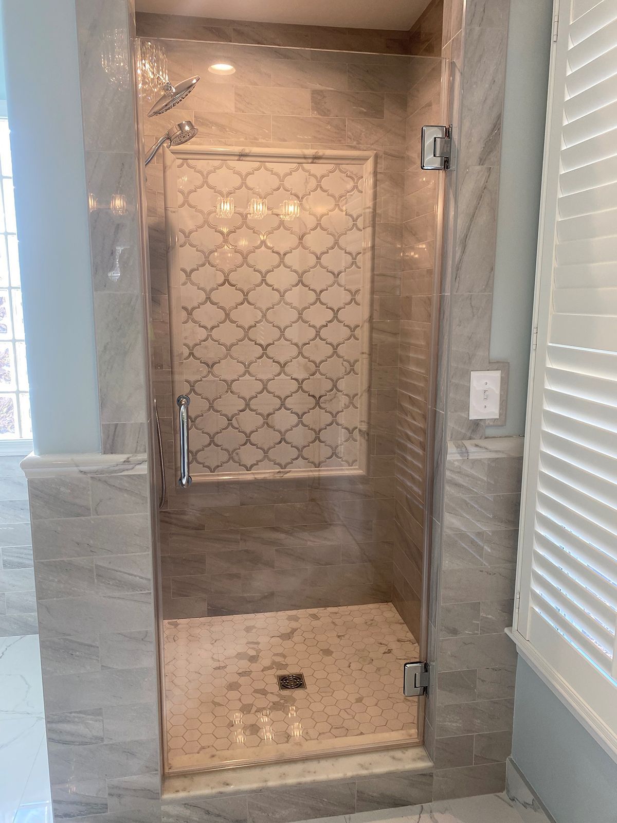 A glass-door shower with beige marbled walls and a decorative, lattice-patterned tile niche.