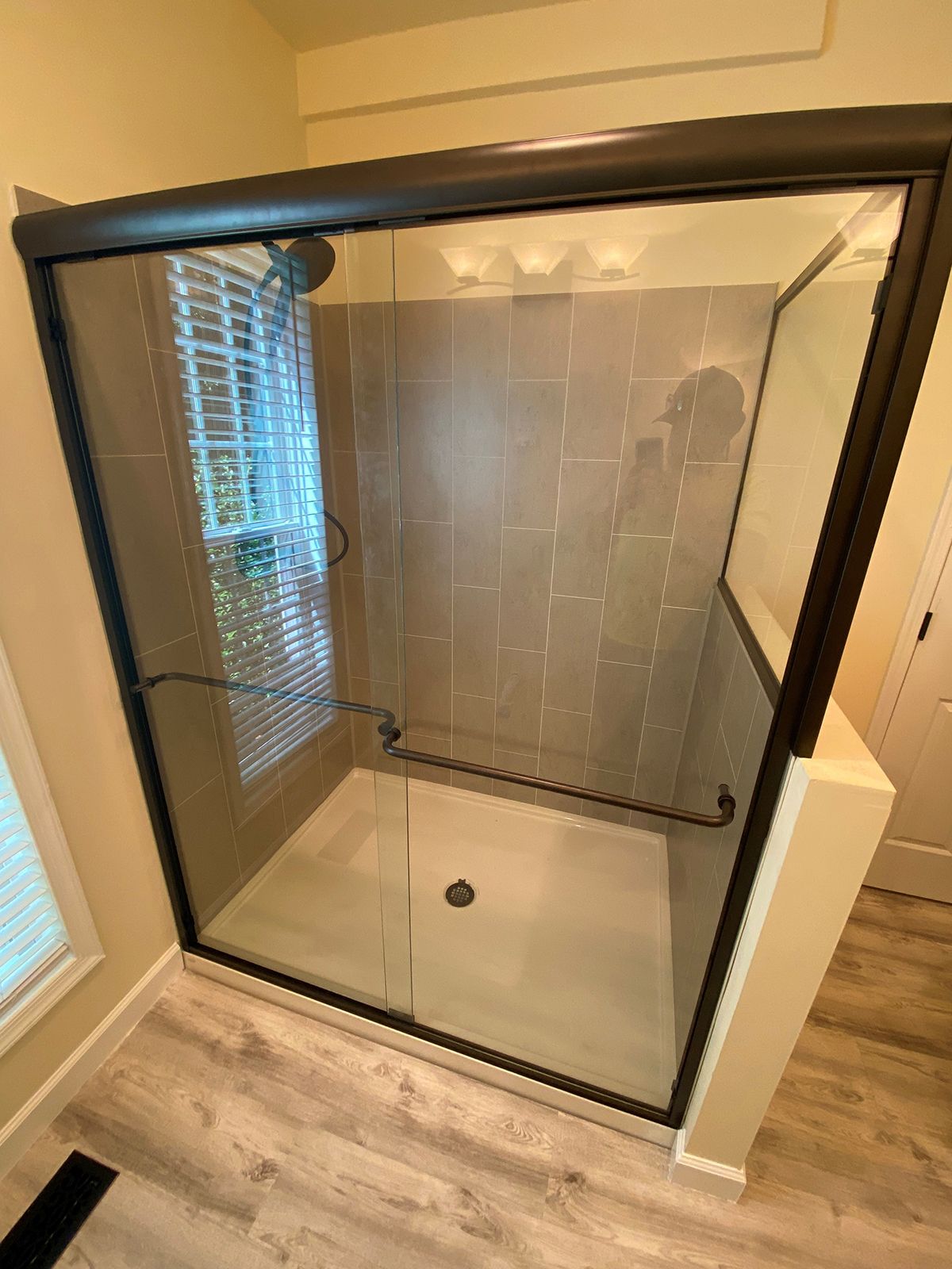 A walk-in shower with a glass sliding door, black frame, gray tiled walls, and light wood-look flooring.