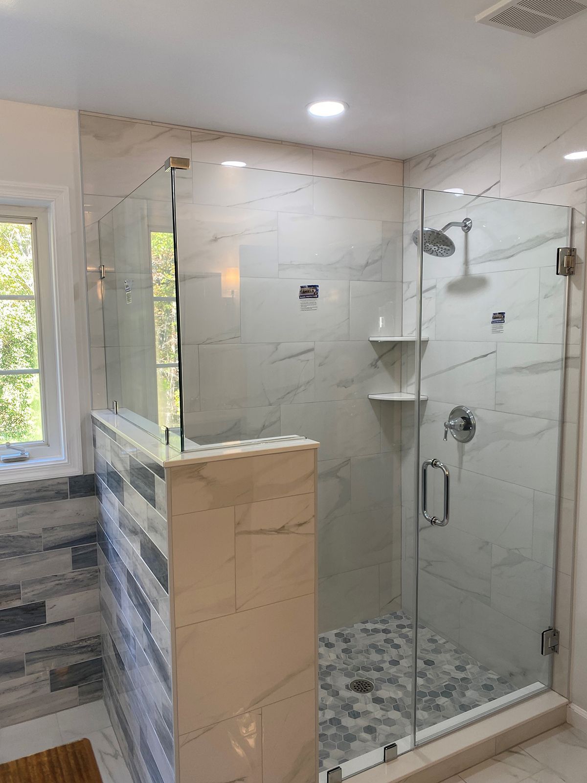 A walk-in shower with marble-look tile, glass door and panel, chrome fixtures, and built-in corner shelves.