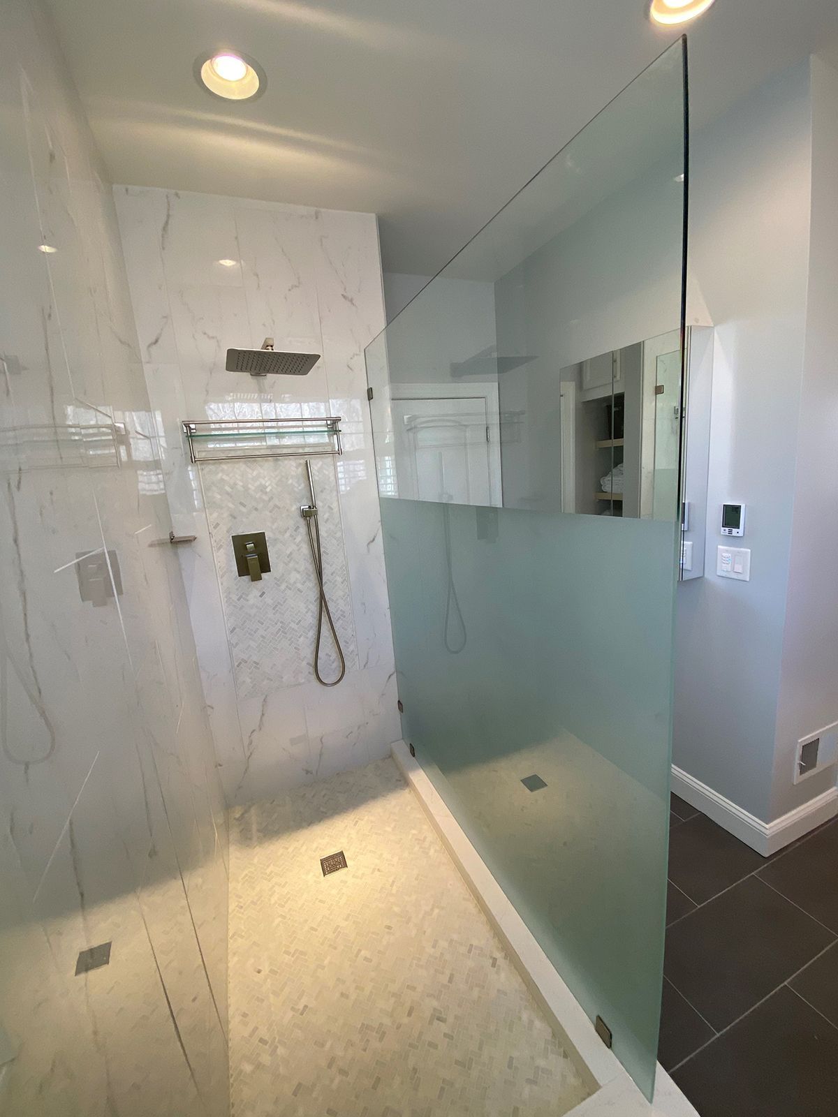 Modern walk-in shower with marble-style walls, a frosted glass panel, rainfall showerhead, and light-colored pebble floor.