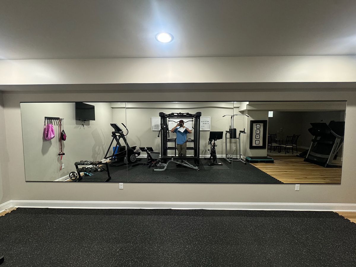 A person stands in a home gym reflecting in a large wall mirror, which shows equipment including a treadmill and rack.