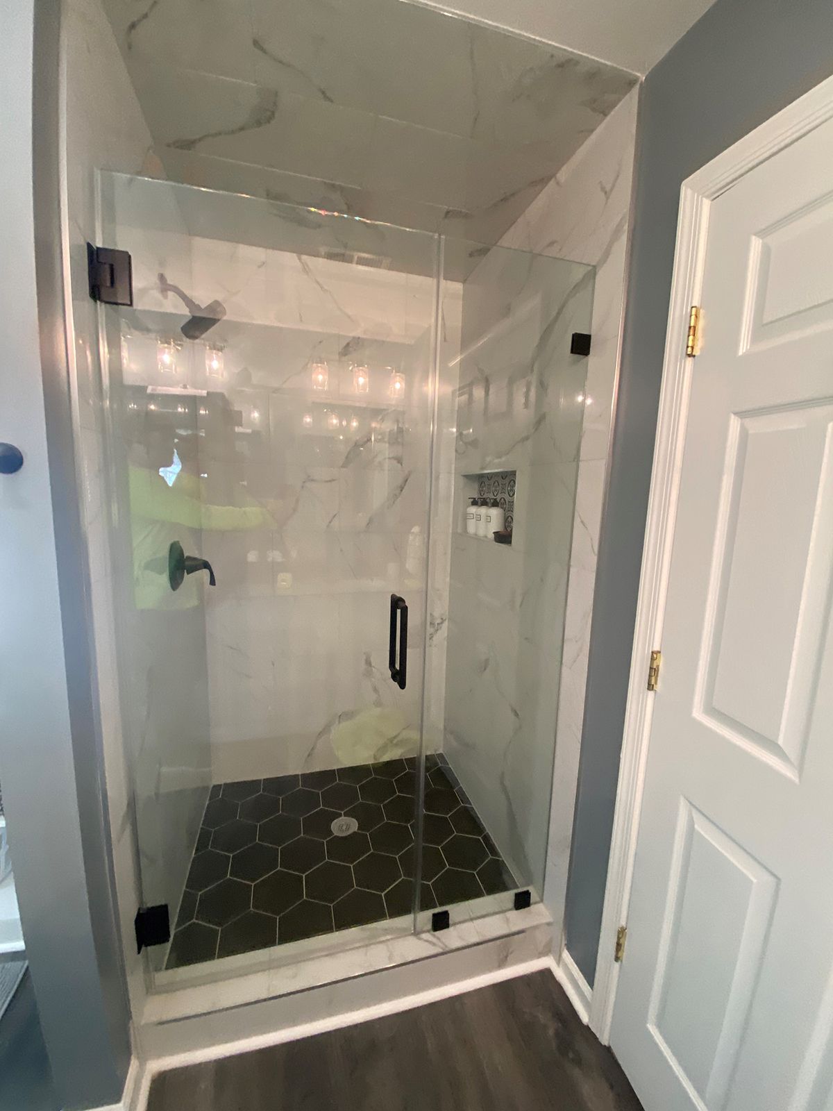 A glass-enclosed shower with white marble-patterned walls and a dark hexagonal tile floor, situated beside a white door.