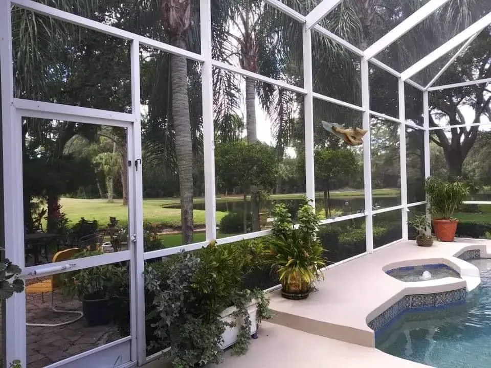 Screened-in patio overlooking a pool and garden; white frame, plants, lush green landscape.