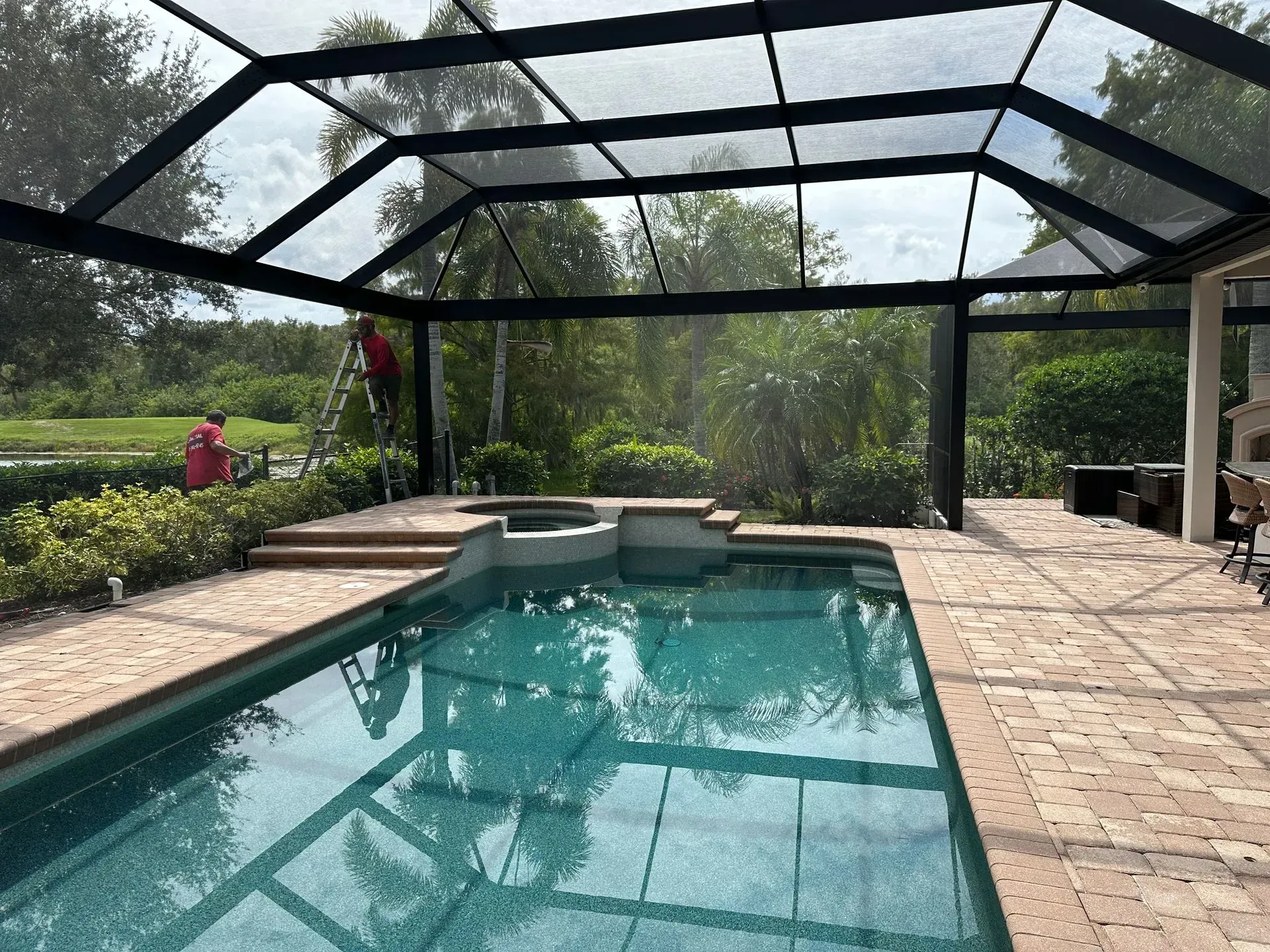 Pool under screened enclosure; dark-framed structure over a pool with surrounding brick; greenery in background.