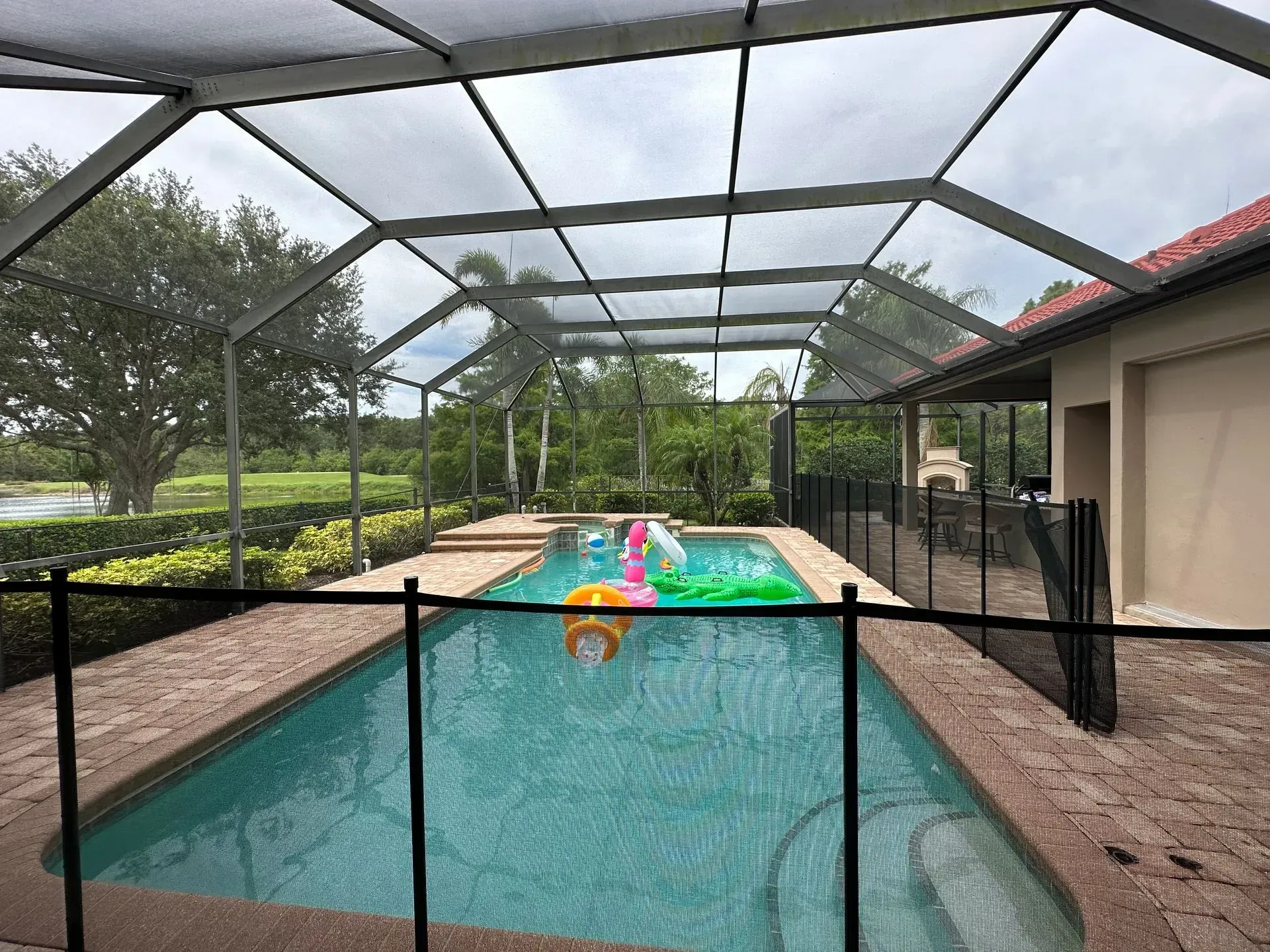 Screened-in pool with inflatable toys; black safety fence, tan patio, and trees in the background.