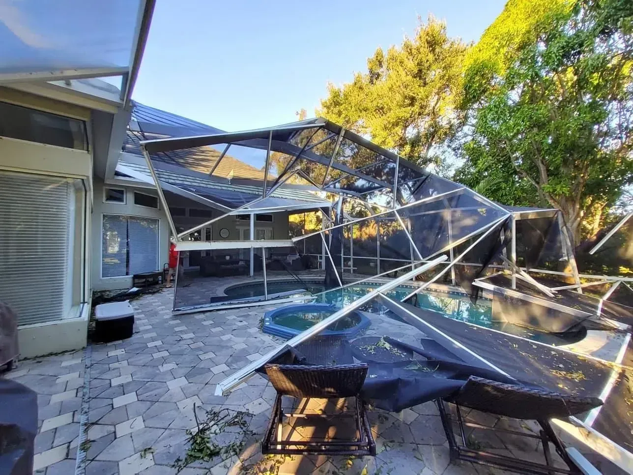 Damaged pool enclosure. Black screen and white frame are collapsed over a pool and patio in daylight.