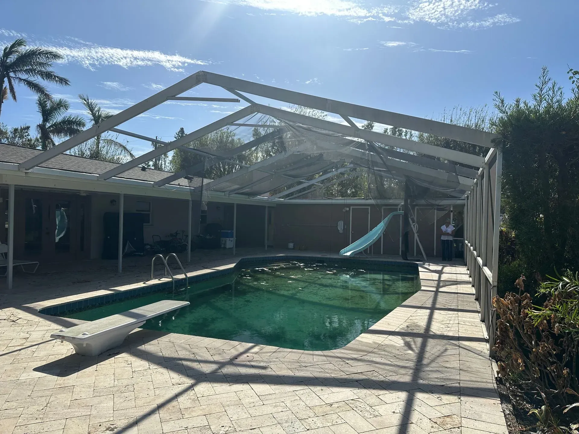 Pool covered by a screened enclosure. Green pool with diving board. Overcast sky.