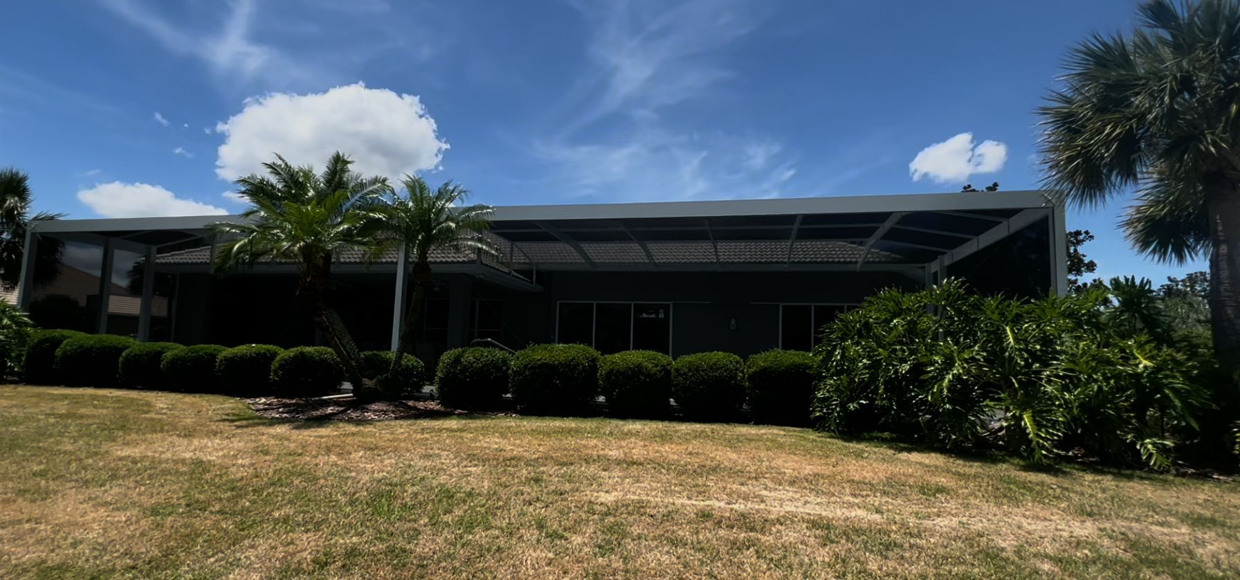 A long, single-story building with a screened porch, surrounded by greenery and a blue sky with clouds.