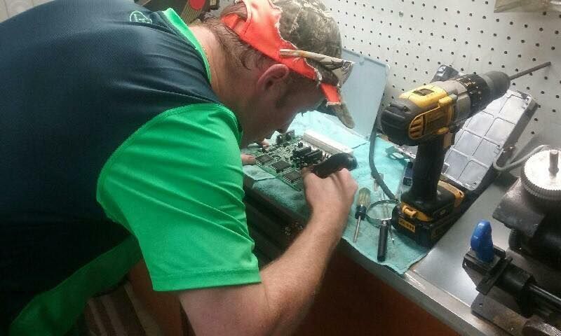 Man works on machinery with a drill, workbench, and tools. He wears a green shirt and camo headgear.