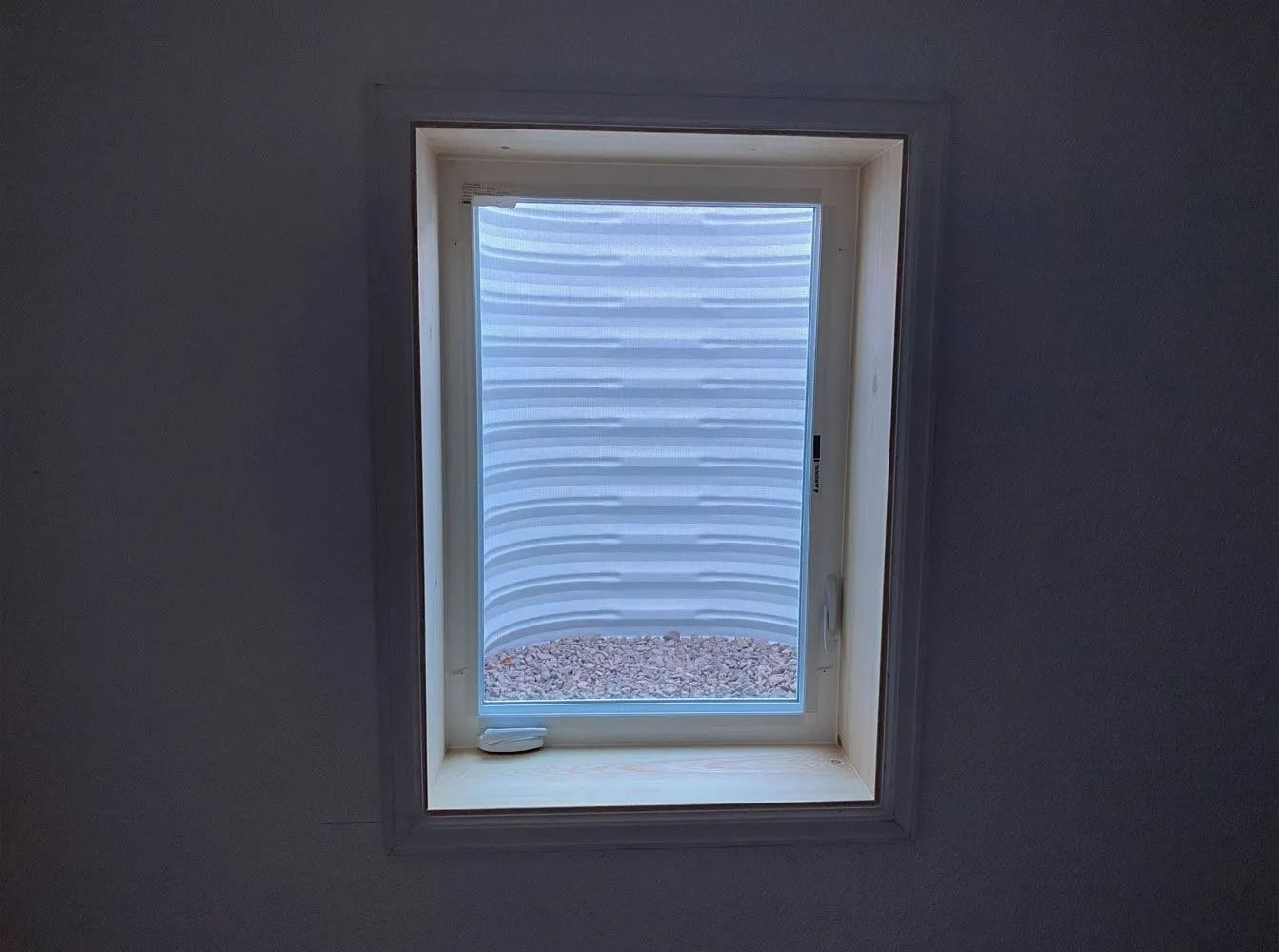 Small rectangular basement window with white trim. Visible through the window is a corrugated well and gravel.
