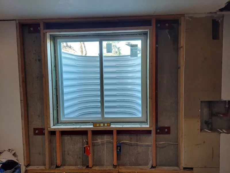 Framed window in basement wall with white blinds, concrete foundation, and exposed framing.
