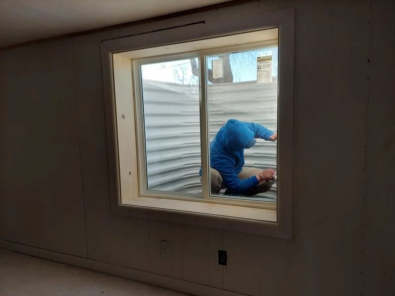 Person in blue bending at a basement window, looking out. Window has white trim, corrugated metal outside.