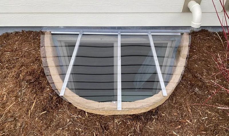 Basement window well with clear cover and metal bars; surrounded by mulch and near a downspout.