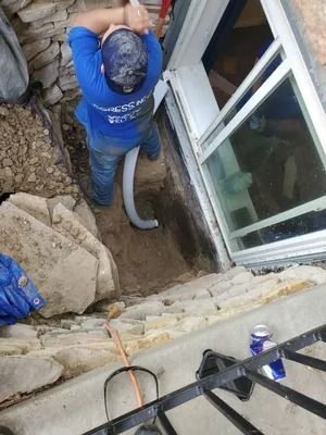 Man in blue shirt works on drainage near a window and stone wall.