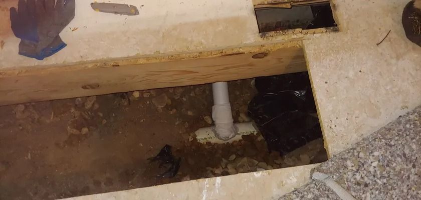 Underground view of a PVC pipe, likely for plumbing, framed by exposed wood and concrete. Dark debris and a glove are visible.