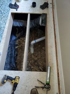 Wooden floor opening showing ductwork, plumbing, and tools like a drill and caulk gun.