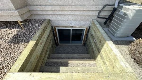 Wooden staircase leading to a basement window well; air conditioning unit in the background.