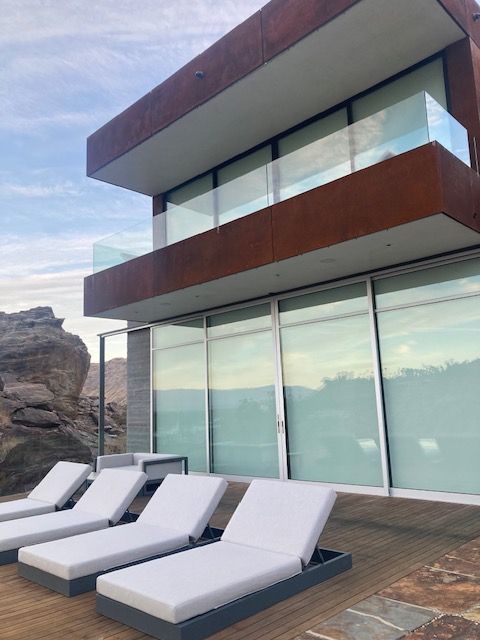 Modern home with a flat roof and glass windows overlooking a rocky landscape, featuring lounge chairs.