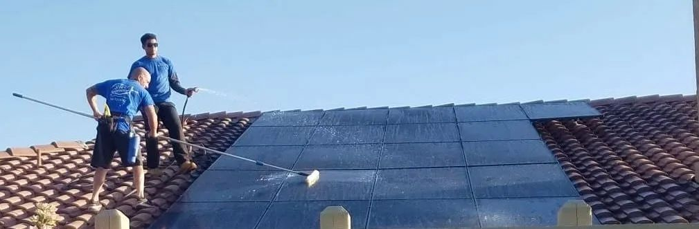 Two men in blue shirts on a tiled roof installing solar panels under a blue sky.