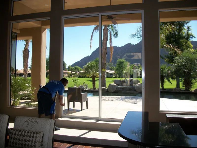 A person cleaning a sliding glass door, overlooking a yard, pool, and mountain range.