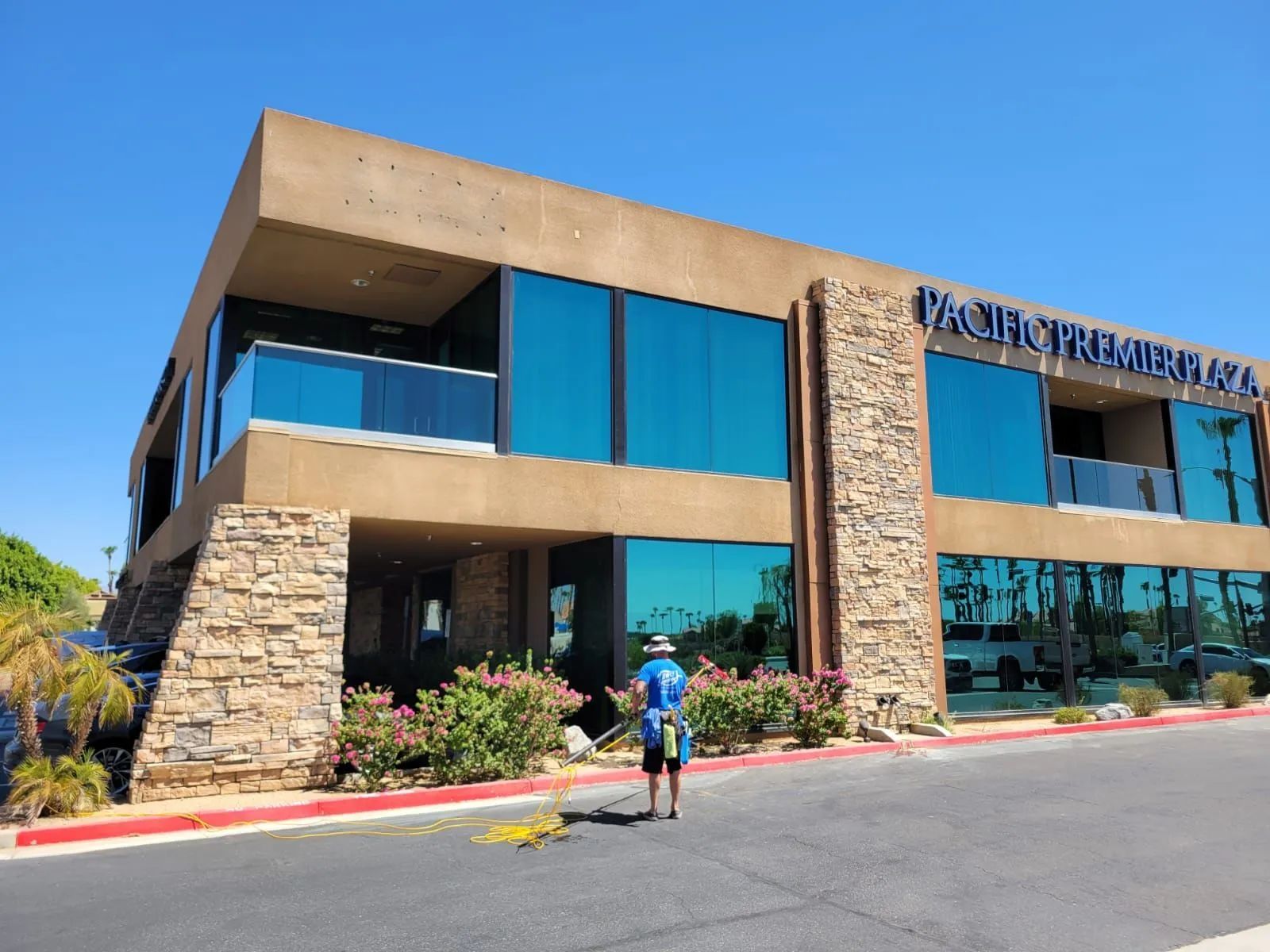 Two-story building with teal windows and stone accents; person wearing blue shirt working in front on a sunny day.