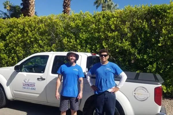 Two men in blue shirts stand next to a white pickup truck in front of green bushes under a sunny sky.