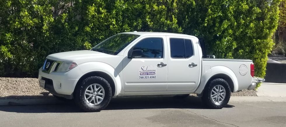White Nissan Frontier pickup truck parked on a street. Green hedge in the background.