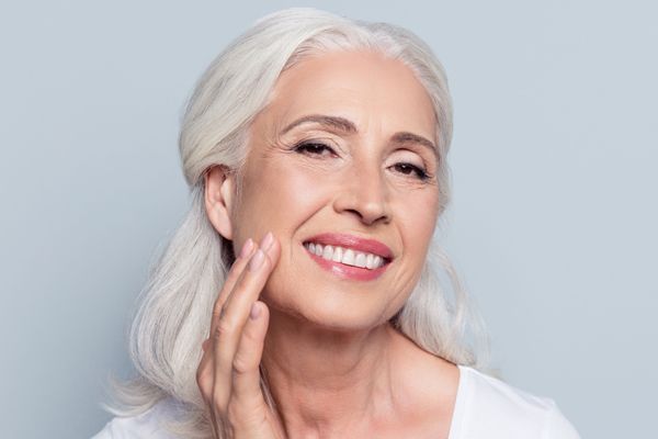 A woman with gray hair is smiling and touching her face.