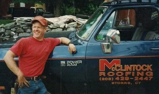 Man in red shirt and cap leaning against dark blue "McClintock Roofing" truck, smiling.
