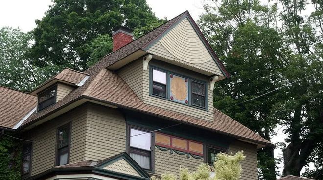 Two-story house with brown shingle roof, green siding, and a gabled dormer. Green trees in the background.