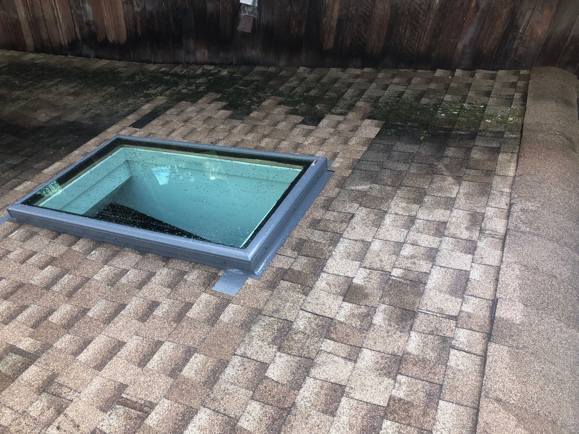 Skylight on a brown shingled roof with green moss, viewed from above.