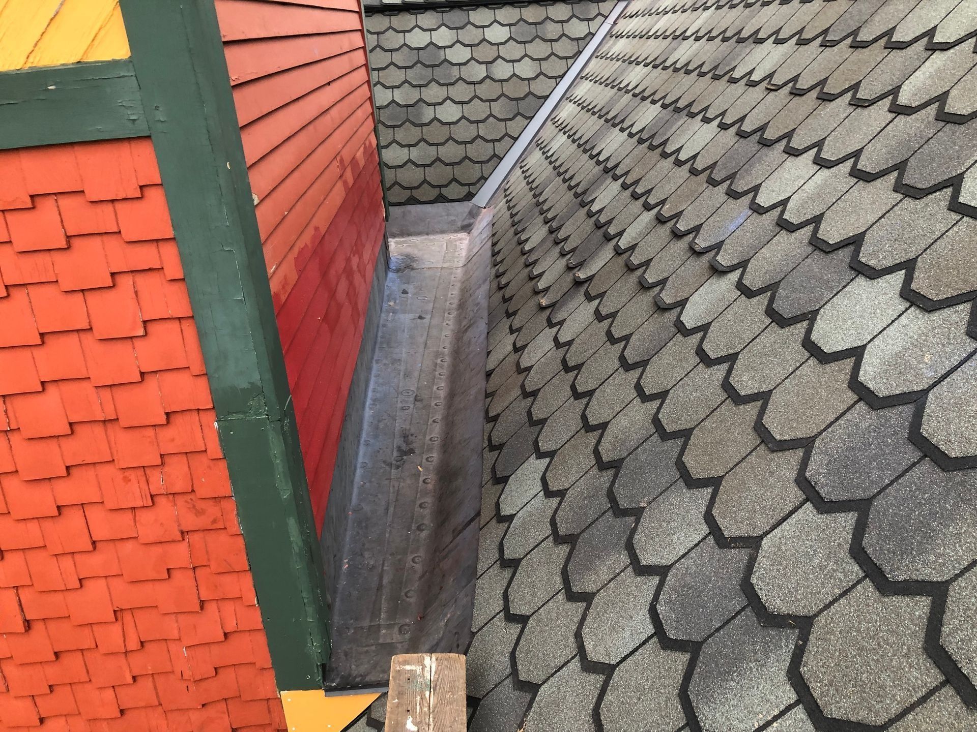 View of a roof valley with overlapping gray shingles on one side and a red wall with orange shingles.