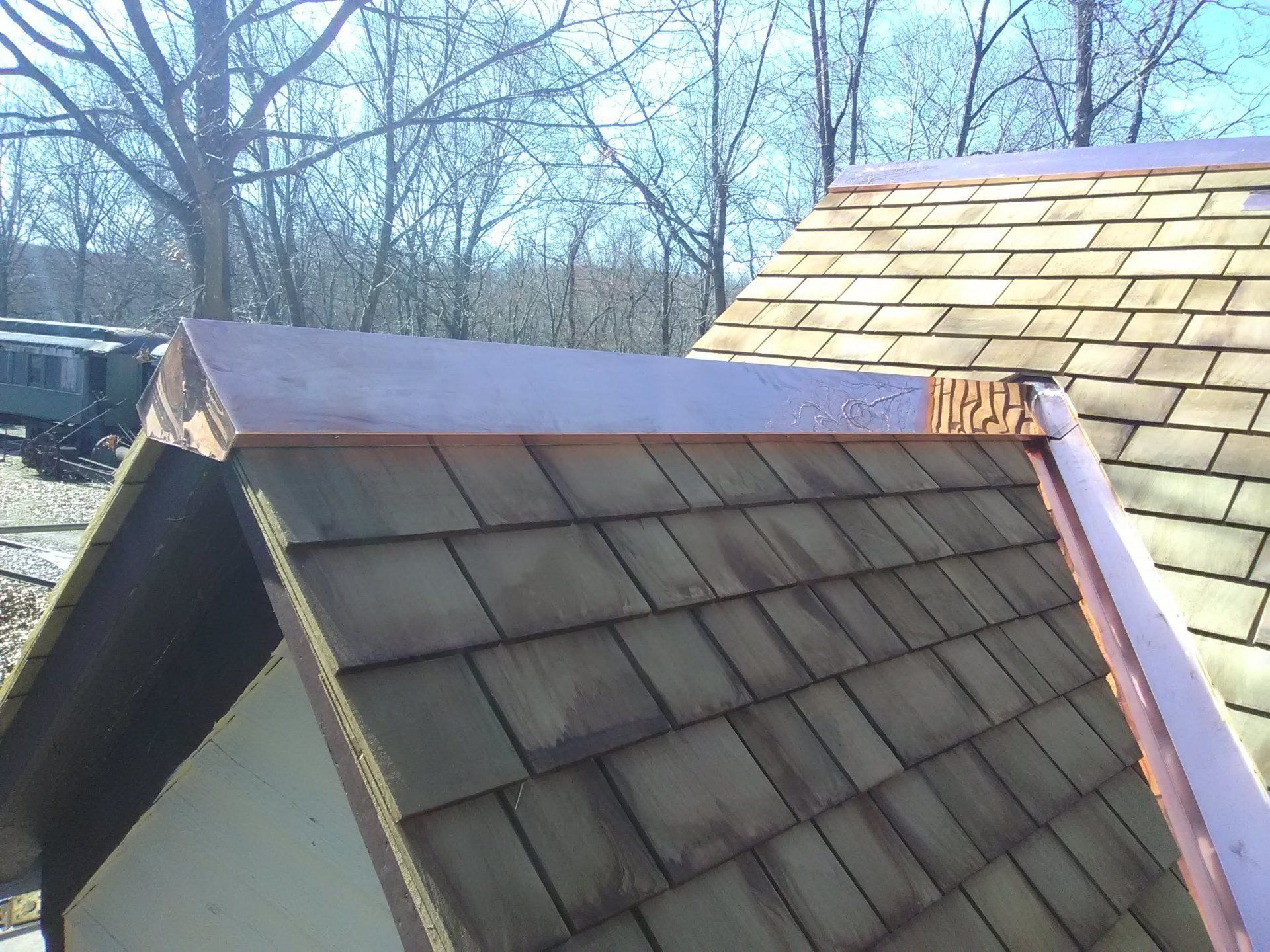 Copper flashing on brown shingle roof, edge detail. Sunlight, trees in background.