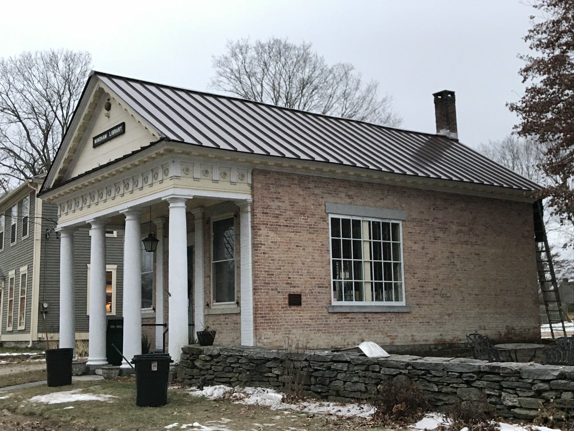Brick building with a columned porch and a stone wall, under a cloudy sky with some snow on the ground.