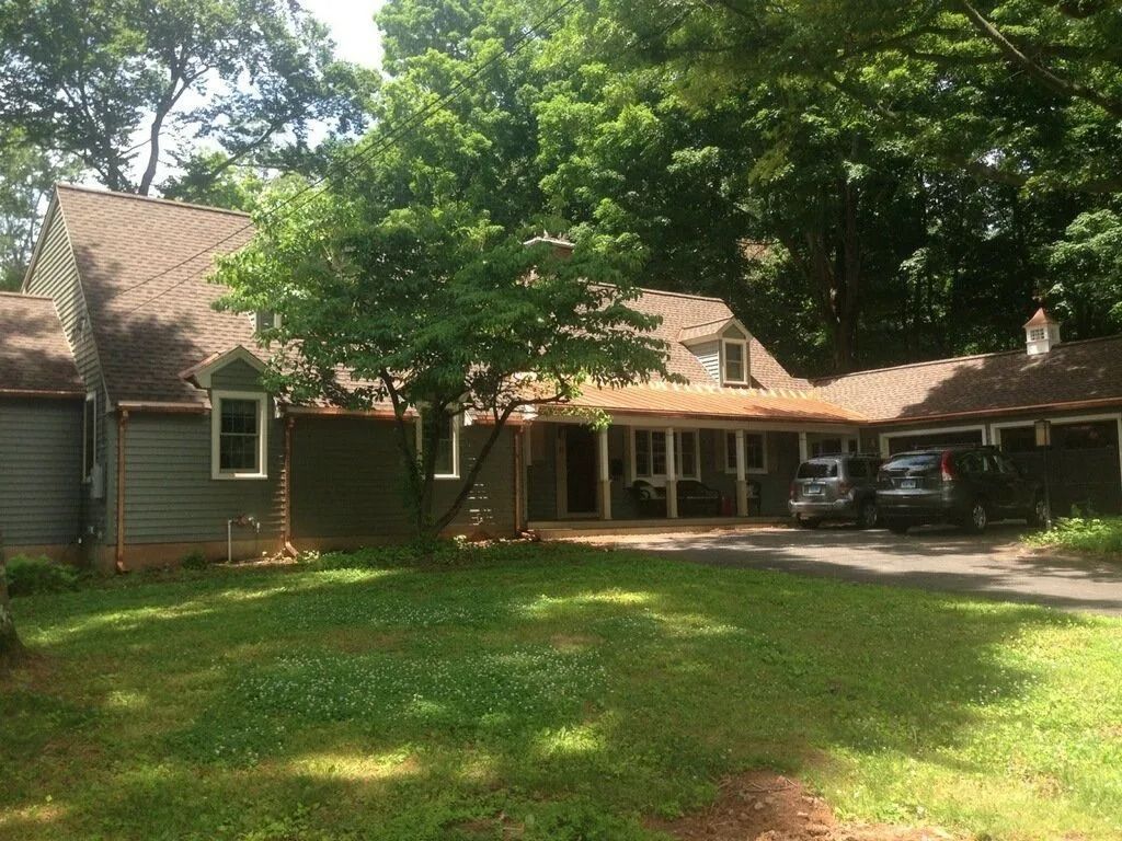 Green house with brown roof and cars parked in a driveway, surrounded by trees and grass.