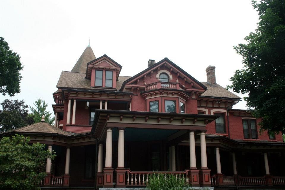 Red Victorian house with a wraparound porch, columns, and a small tower.
