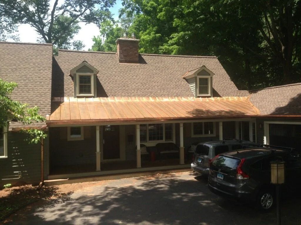 A brown-roofed house with a copper awning. Two cars are parked in front of the house. Trees are in the background.