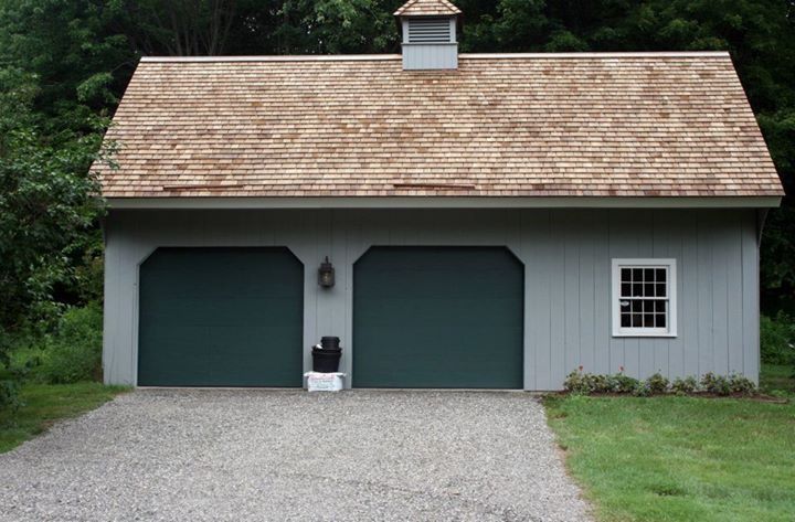 Two-car garage with gray siding, green doors, and a wood shingle roof. A gravel driveway leads up to it.